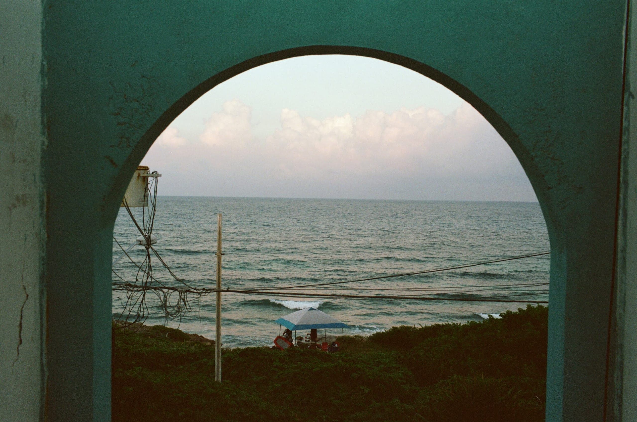 View through a teal archway to the ocean with a beach umbrella and tangled wires