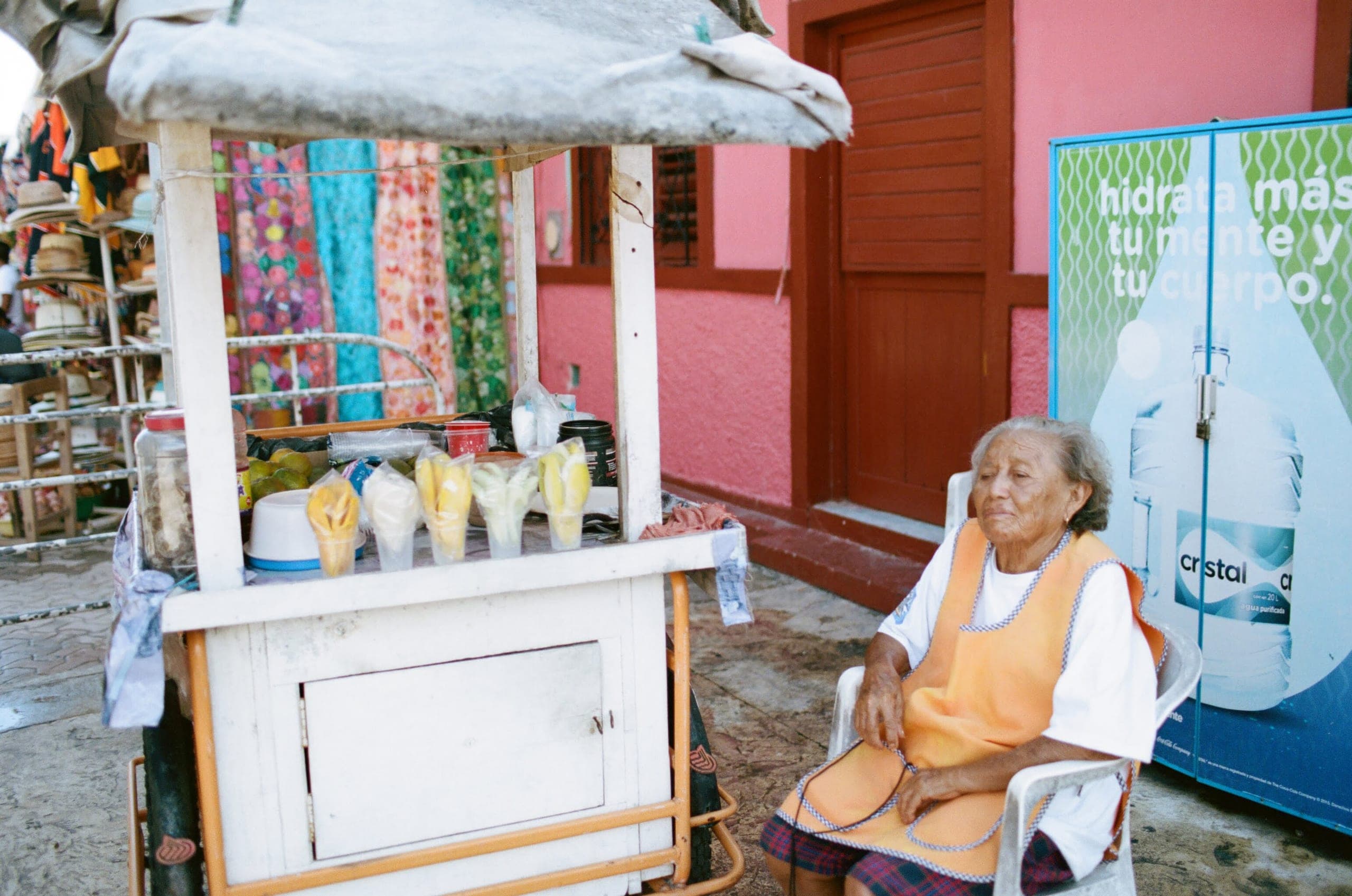 Elderly woman in an orange apron sitting beside a fruit cart in front of a pink wall