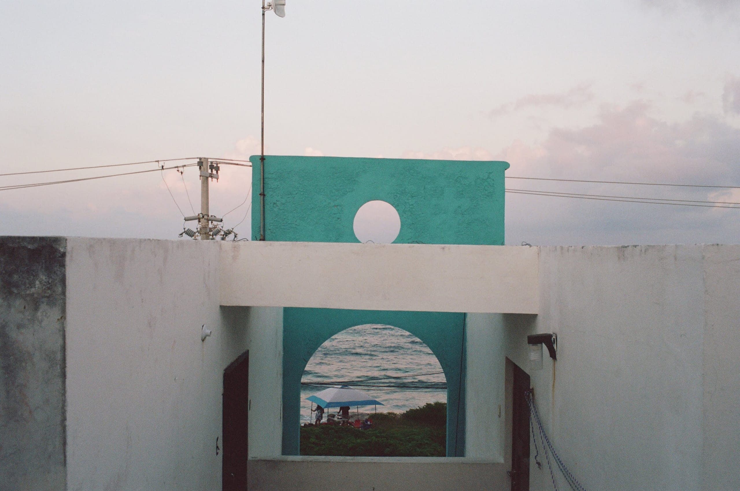 White and teal building with a circular cutout above an archway that frames the ocean and a beach umbrella