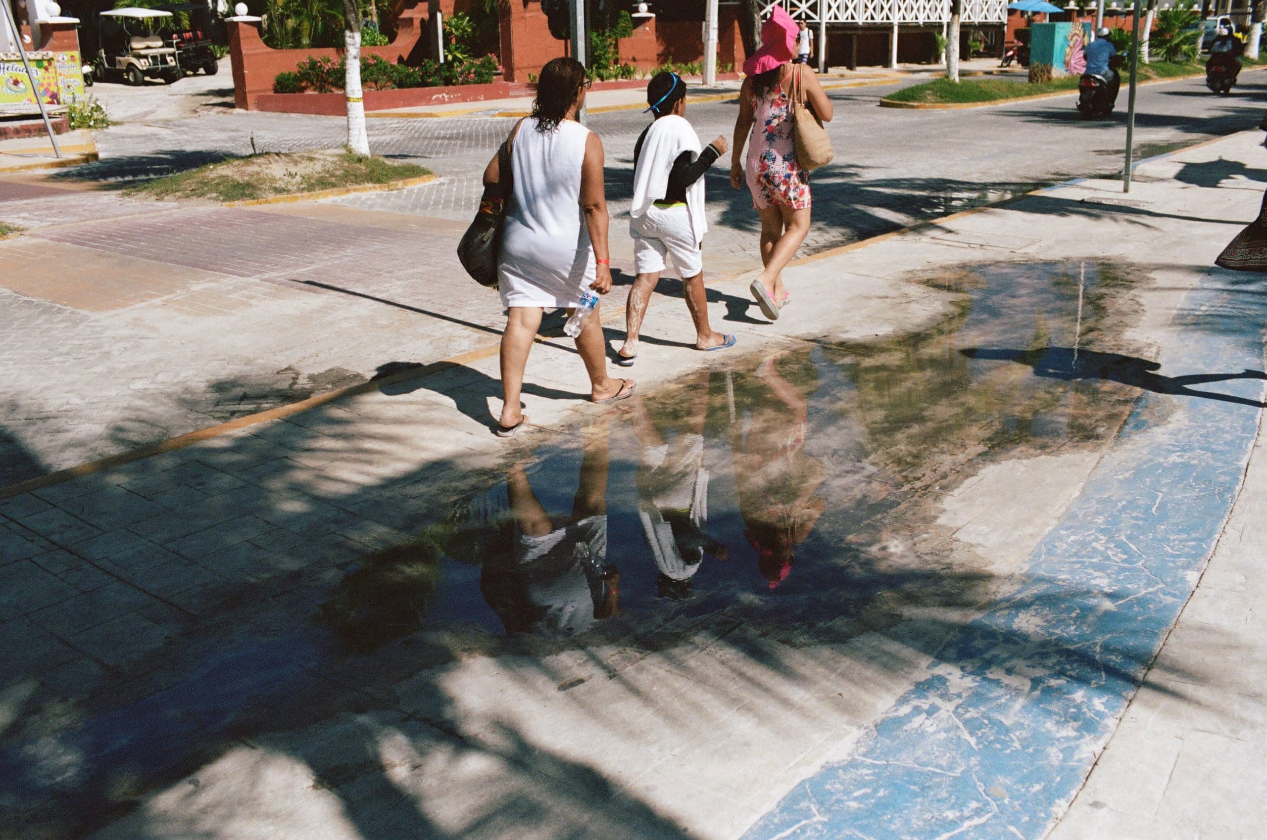 Three people walking past a puddle on a sunny sidewalk, their reflections caught in the water