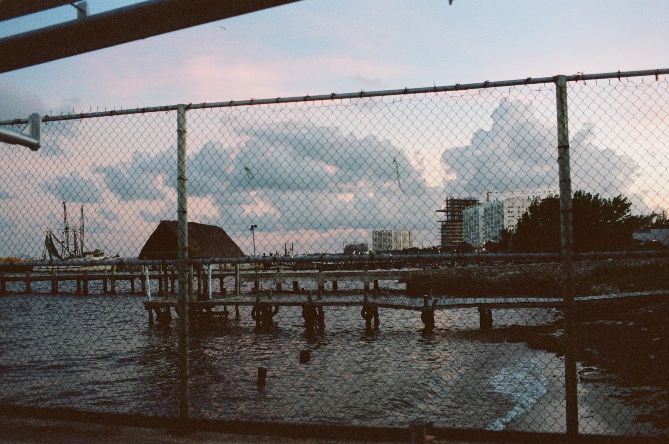 Shrimp boat and palapa hut across water at dusk, viewed through a chainlink fence