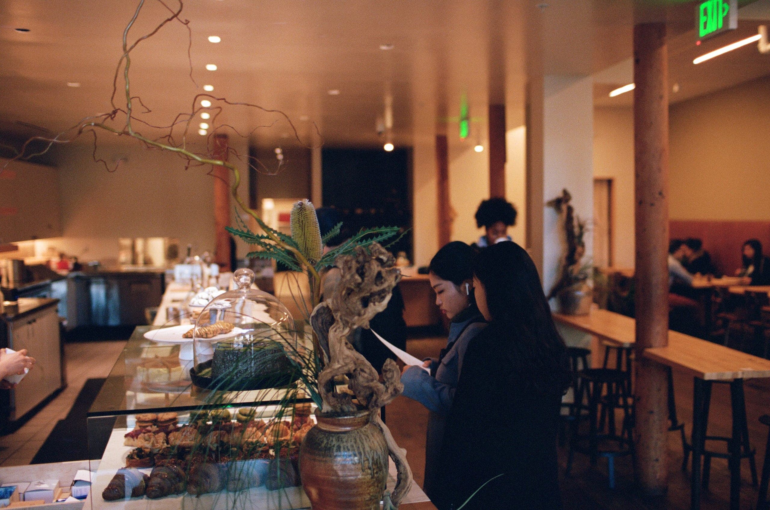 Bakery pastry case with people browsing, wood columns, banksia in a vase